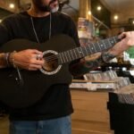 Person playing a small dark-bodied acoustic guitar in a record shop, illustrating the compact scale of a half-size travel guitar