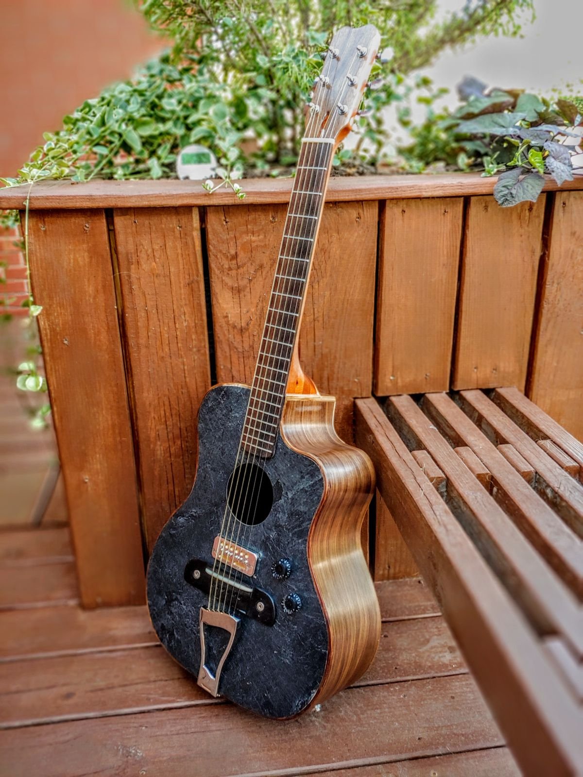 Acoustic guitar with a dark composite top and wood-grain back leaning against a wooden fence outdoors