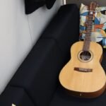 Compact-bodied acoustic guitar resting on a sofa, illustrating the half-size dreadnought shape of a mini travel acoustic