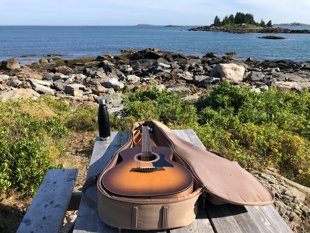 Acoustic guitar resting in its open padded travel bag on a coastal picnic table