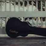 A guitar case resting against a white fence on a pavement, waiting to travel