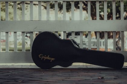 A guitar case resting against a white fence on a pavement, waiting to travel