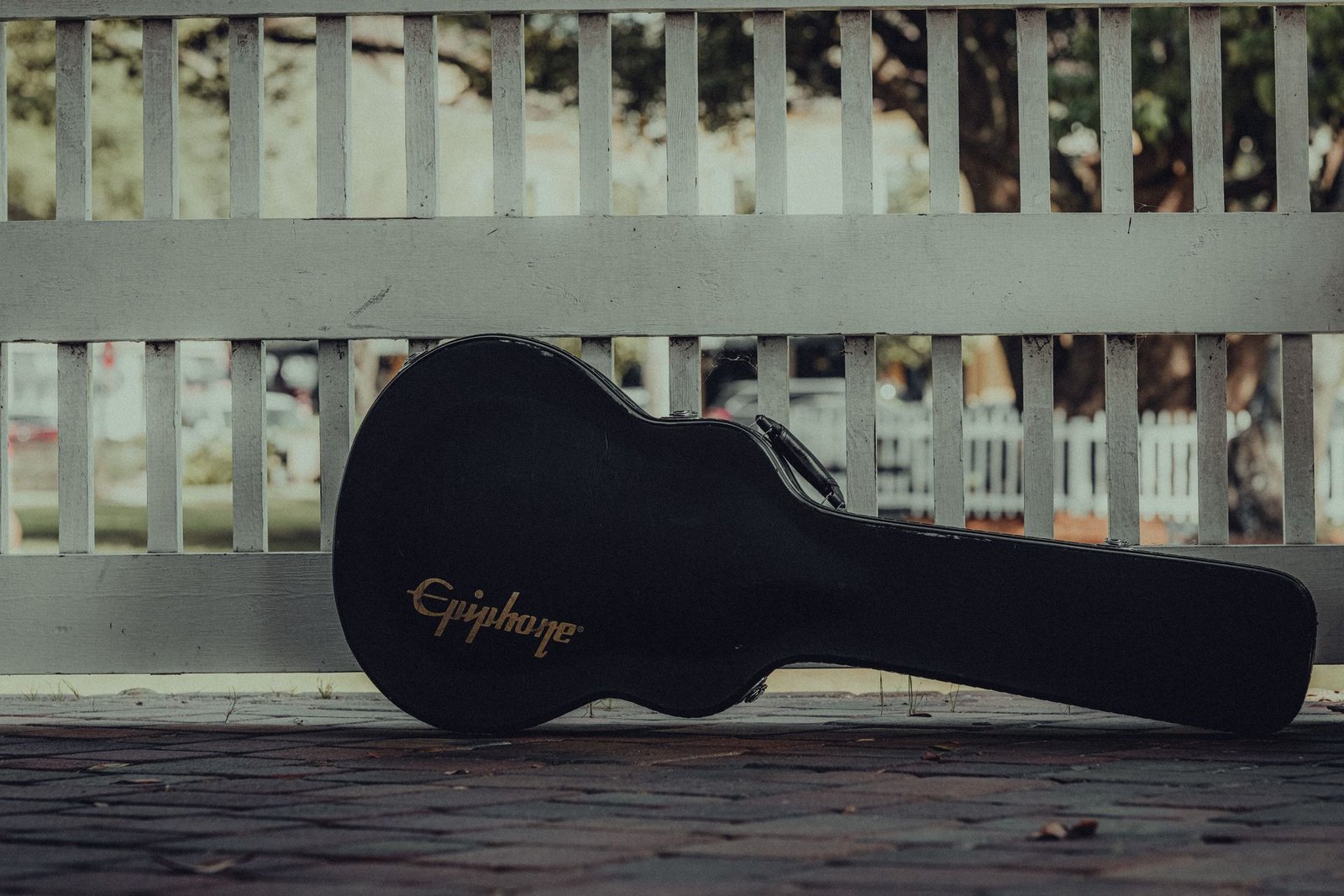 A guitar case resting against a white fence on a pavement, waiting to travel