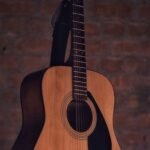 Natural-finish steel-string acoustic guitar hanging by its strap against a brick wall, showing the full body and neck