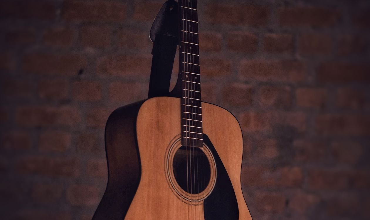 Natural-finish steel-string acoustic guitar hanging by its strap against a brick wall, showing the full body and neck