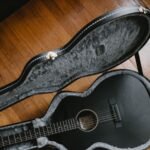 A matte black acoustic guitar resting in an open hardshell case with plush grey lining, illustrating a travel guitar kit