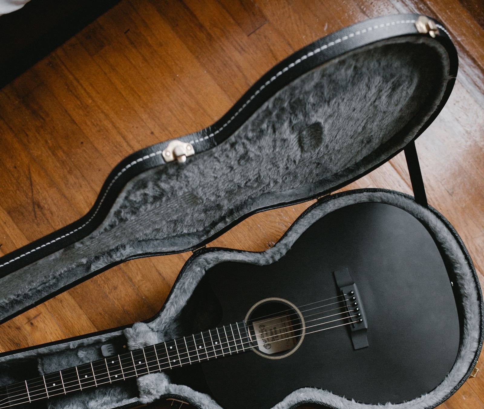 A matte black acoustic guitar resting in an open hardshell case with plush grey lining, illustrating a travel guitar kit