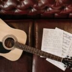 Small-bodied acoustic guitar with sheet music on a leather sofa, illustrating a compact travel-friendly acoustic