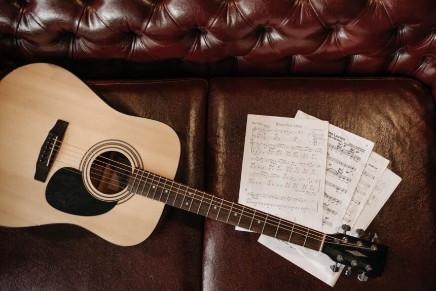 Small-bodied acoustic guitar with sheet music on a leather sofa, illustrating a compact travel-friendly acoustic