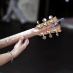 A hand tuning the gold machine heads on a wood-bodied travel acoustic guitar headstock