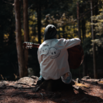 A guitarist sitting on a log playing an acoustic guitar in a forest, the kind of setting a travel guitar for camping needs to handle