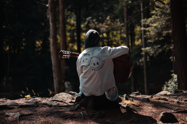 A guitarist sitting on a log playing an acoustic guitar in a forest, the kind of setting a travel guitar for camping needs to handle
