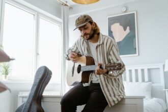 A man tuning an acoustic guitar while sitting on the edge of a bed, with an open guitar case beside him