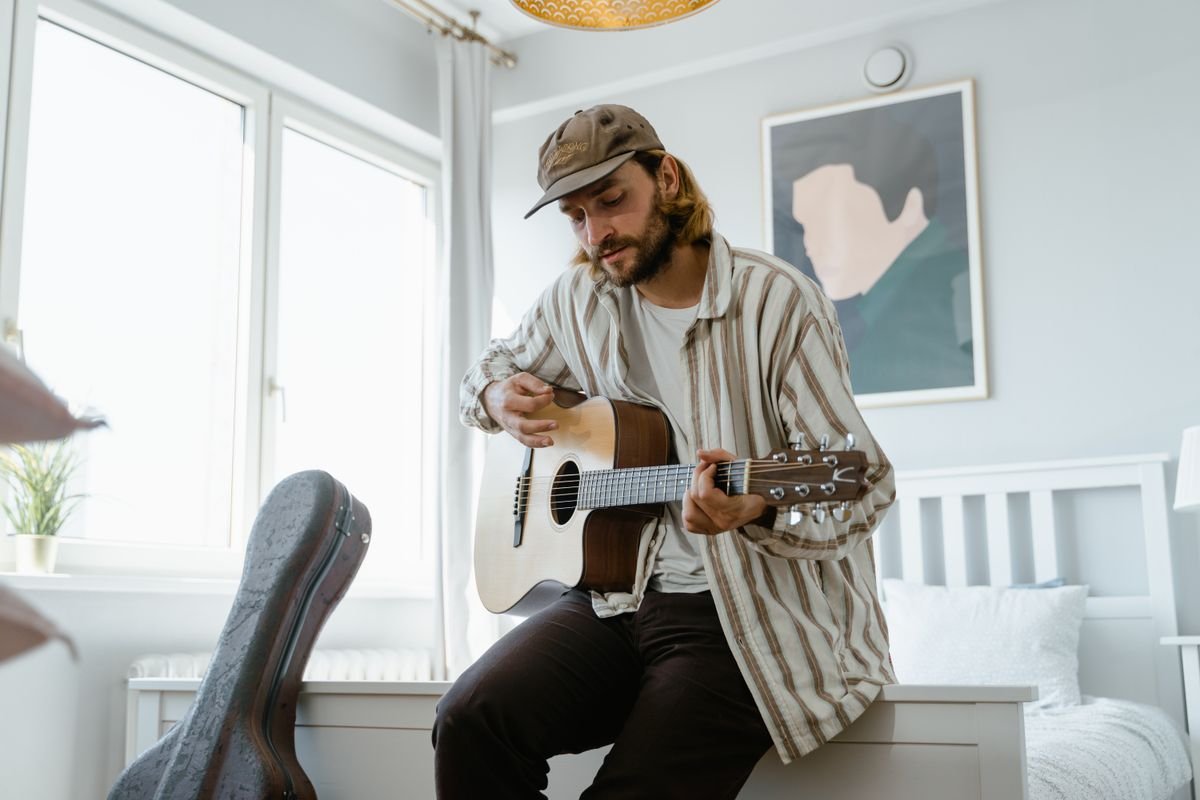 A man tuning an acoustic guitar while sitting on the edge of a bed, with an open guitar case beside him