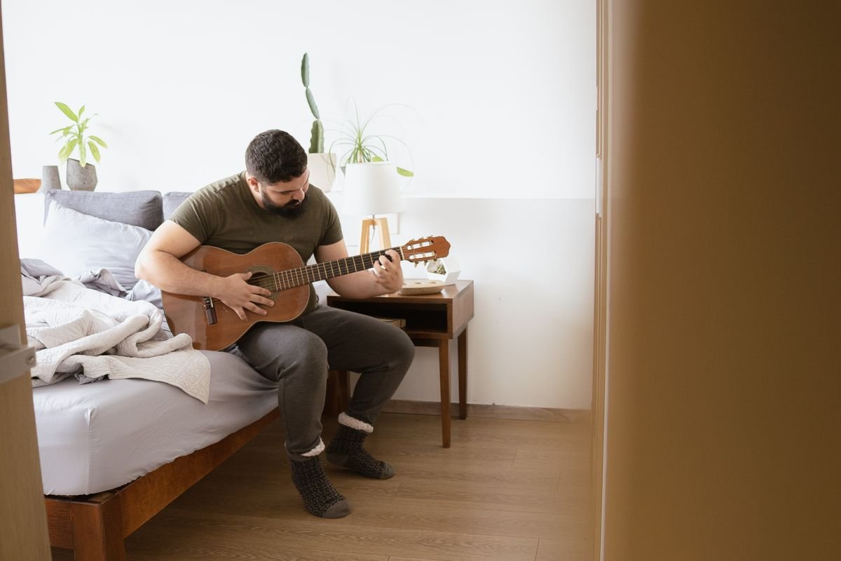 A guitarist practising quietly while sitting on the edge of a bed in a small bedroom