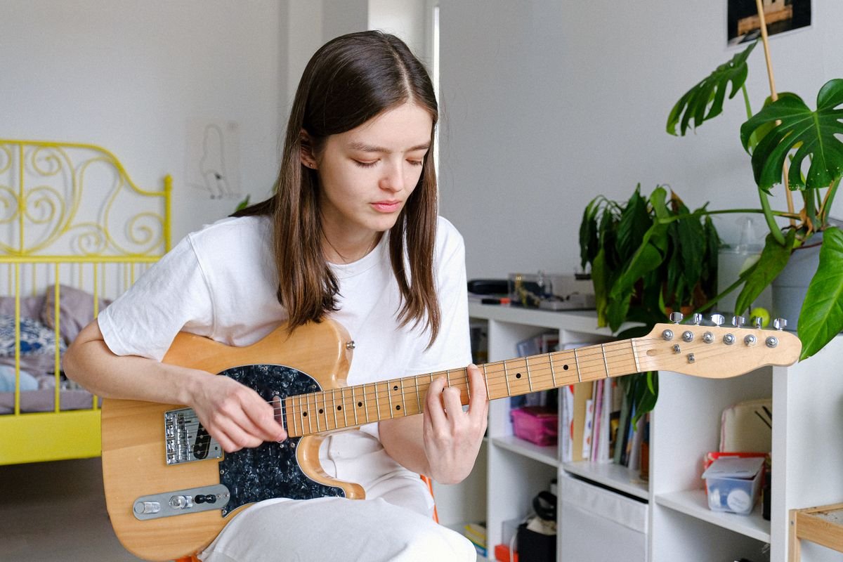 A woman playing an electric guitar in a small, plant-filled bedroom